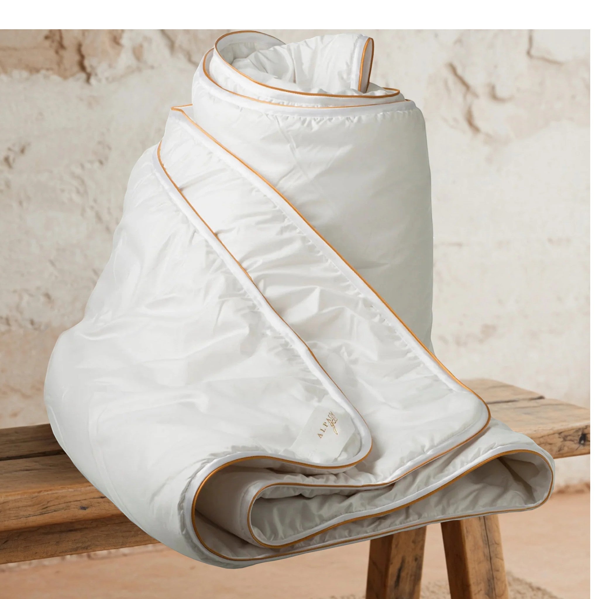 Folded white quilt with gold trim on a wooden surface against a beige wall.