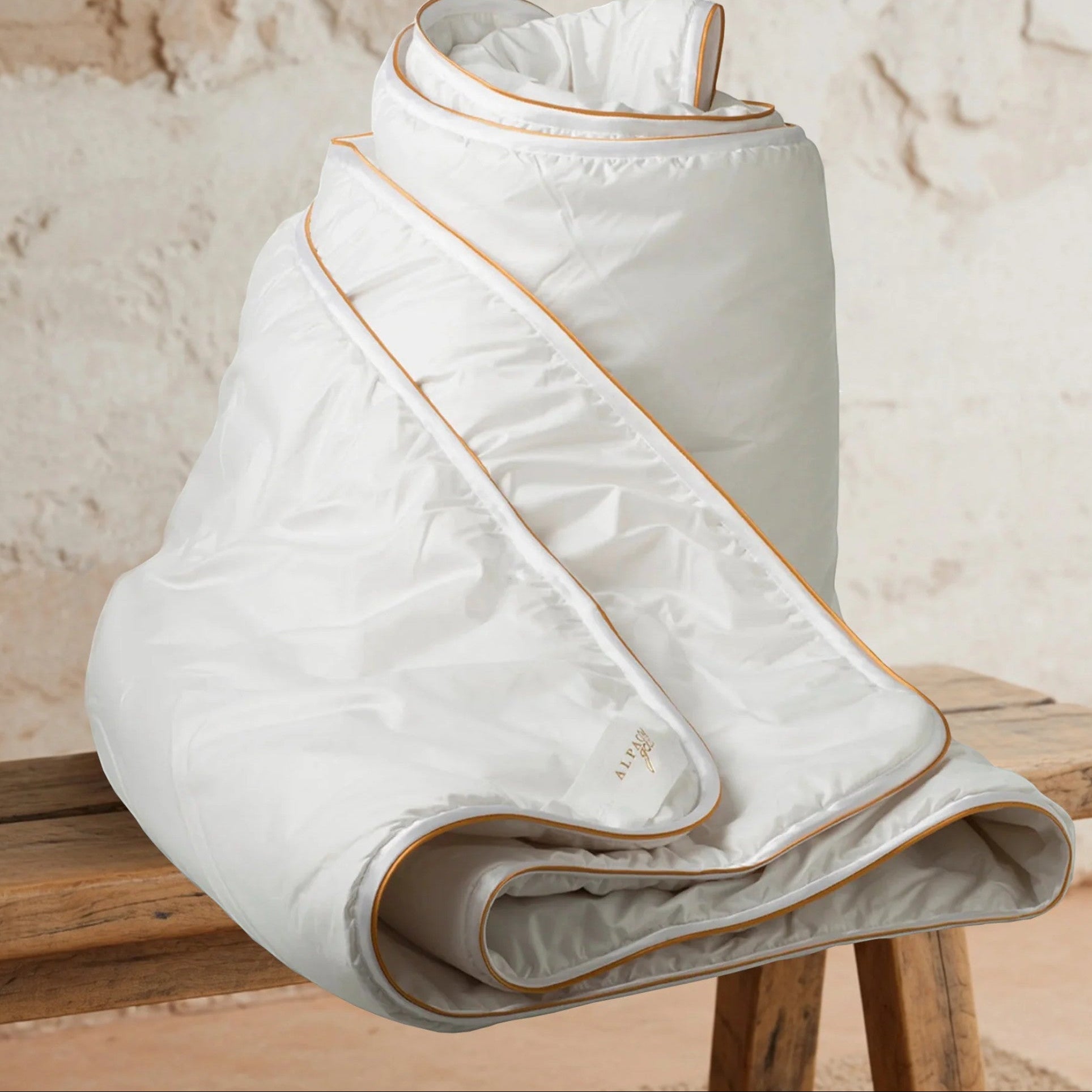 Folded white quilt with gold trim on a wooden surface against a beige wall.