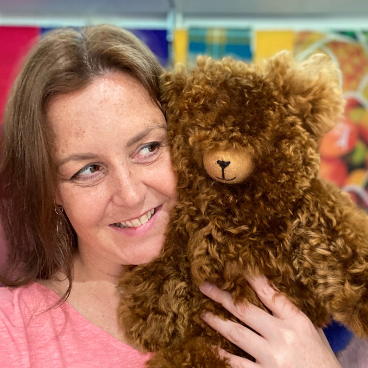 A smiling woman with light brown hair holds a large, curly brown teddy bear close to her face.
