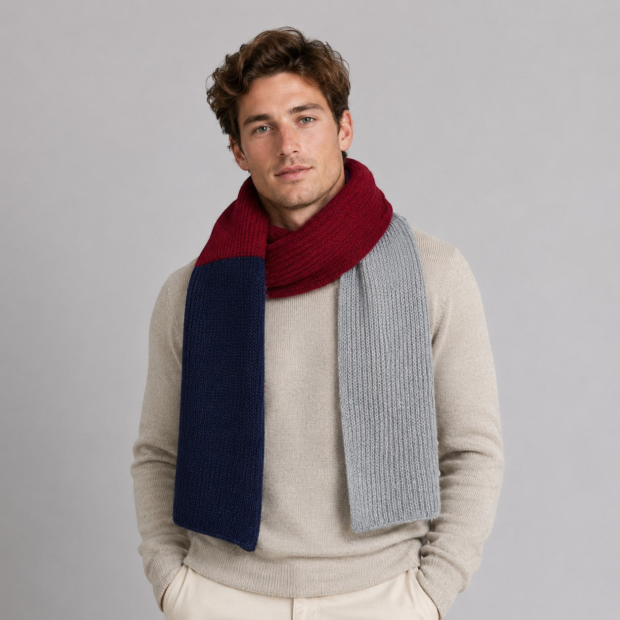 A man wearing a thick knit colourblock scarf in navy blue, burgundy red, and light gray, styled over a beige sweater against a solid gray studio background.