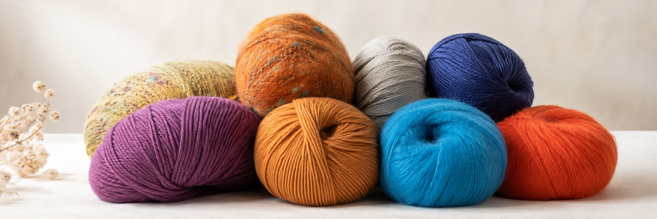 A group of colorful yarn balls in purple, mustard, orange, gray, navy, blue, and red, arranged on a light surface against a soft beige background.